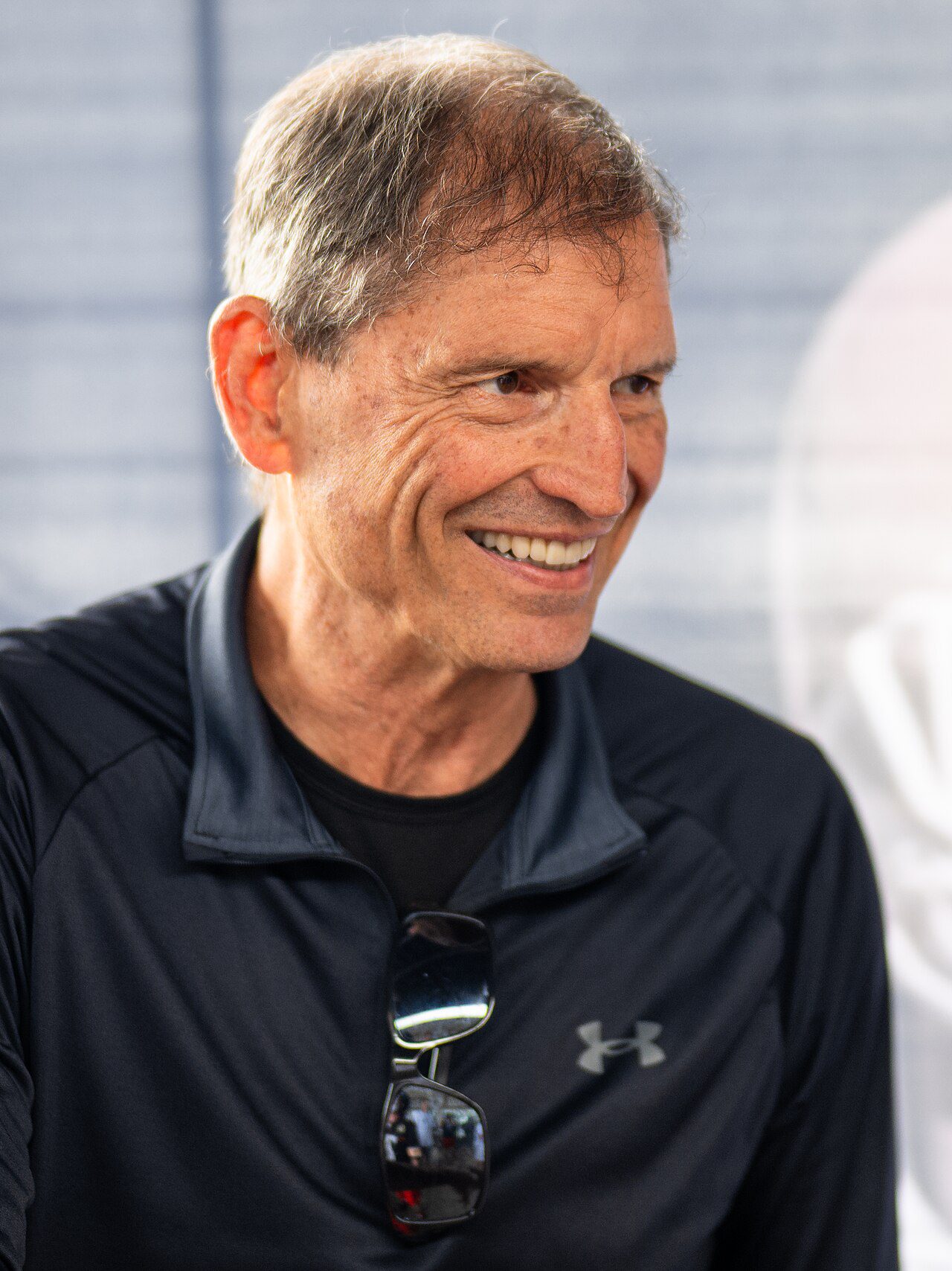 Bernie Kosar speaking at a public event in a dark jacket and cap.